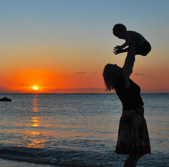 in riva al mare, sullo sfondo di un tramonto, una mamma solleva in alto il suo bambino 